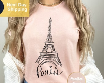 May include: A heather peach t-shirt featuring a black line-art illustration of the Eiffel Tower centered on the chest. Below the tower, the word Paris is written in a black cursive script with a small heart icon above the letter i.