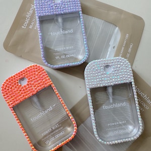 May include: Three clear, rectangular hand sanitizer spray bottles with rhinestone embellishments. The bottles are in shades of orange, white, and lavender. Each bottle has the text "touchland" and "POWER MIST" printed on it.