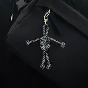 Puede incluir: Un llavero de cuerda en blanco y negro con forma de persona. El llavero está sujeto a un mosquetón plateado. El fondo es una mochila negra.