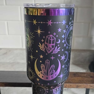 May include: A black stainless steel tumbler with a rainbow iridescent finish. The tumbler features a design of moons, stars, crystals, and other celestial elements.