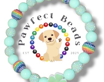 May include: A light green beaded bracelet with rainbow and glitter accents. The bracelet has a four-leaf clover charm and the text "Pawfect Beads Est 2023".