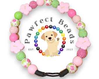 May include: A pink and green beaded dog collar with flower charms. The collar has a black adjustable buckle. The collar is decorated with pink, green, and white beads. The beads are made of plastic and have a glossy finish. The flower charms are made of wood and have a pink paint finish. The collar is designed for small dogs. The collar is labeled "Pawfect Beads Est. 2023".