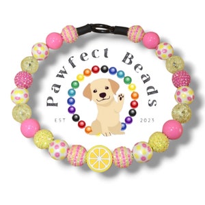 Könnte beinhalten: Ein gelbes und pinkfarbenes Perlenhalsband für Hunde mit einem Zitronenanhänger. Das Halsband besteht aus verschiedenen Perlen, darunter Glitzer-, Tupfen- und Streifenperlen. Das Halsband ist mit dem Text "Pawfect Beads Est 2023" verziert.