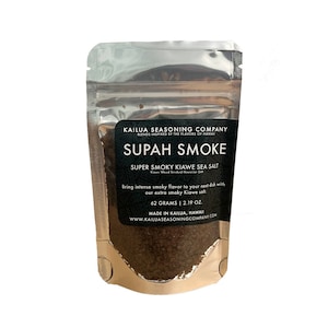 Puede incluir: Una bolsa plateada de sal marina "Supah Smoke" súper ahumada Kiawe. La etiqueta muestra el logotipo de Kailua Seasoning Company y la información del producto. La bolsa contiene 62 gramos de sal, ideal para añadir sabor ahumado a los platos.