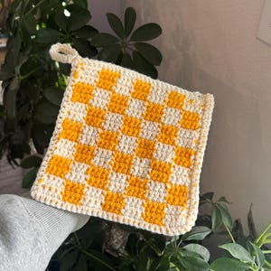 May include: A square, handmade crochet potholder with a yellow and white checkerboard pattern. The potholder has a loop for hanging and is surrounded by green leafy plants.