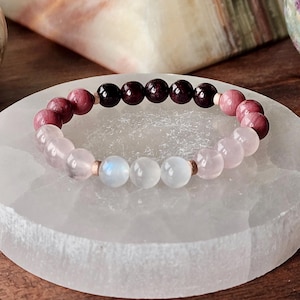 May include: A beaded bracelet featuring a mix of gemstones. The bracelet includes pink, white, and dark red beads. The bracelet is resting on a white, round surface. The bracelet is a piece of jewelry.