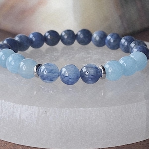 May include: A beaded bracelet featuring alternating light blue and dark blue gemstone beads. The bracelet is displayed on a white, rounded rectangular surface. The beads are round and appear to be polished.