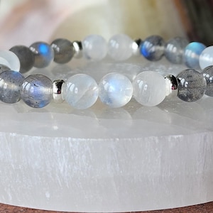 May include: A beaded bracelet featuring alternating gray and white gemstone beads with silver accents. The bracelet is displayed on a white, rectangular surface. The gray beads have blue and iridescent flashes.