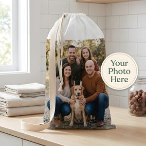 May include: A white drawstring laundry bag with a family photo printed on the front. The photo shows a family of five, including a dog. The bag has a beige strap and the words "Your Photo Here" on a circular sticker.