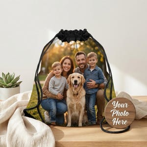 May include: A black drawstring backpack featuring a family portrait with a golden retriever. A wooden round tag with the text "Your Photo Here" hangs from the bag. The background shows a sunny outdoor scene.