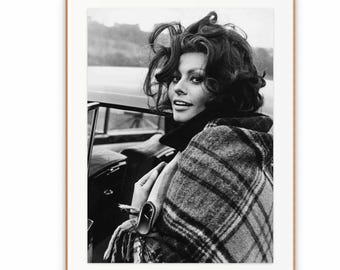 May include: A black and white photographic print featuring a person with voluminous dark hair, wearing a plaid patterned wrap or shawl over a dark collared garment. The subject is positioned inside a vehicle, looking back toward the camera.