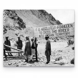 Pode incluir: Uma fotografia a preto e branco mostrando quatro pessoas perto de uma placa de madeira com a inscrição IT IS ABSOLUTELY FORBIDDEN TO CROSS THIS BORDER INTO AFGHAN TERRITORY. Os indivíduos usam túnicas e toucados tradicionais num terreno rochoso.