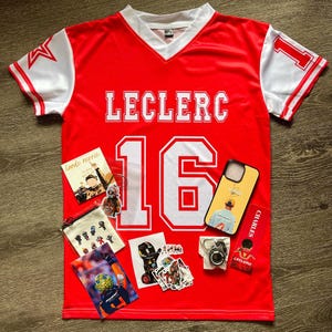 May include: Red and white Leclerc jersey with the number 16. The jersey has white sleeves with a star on one and the number 1 on the other. Several accessories are arranged around the jersey, including a phone case, stickers, and a small pouch.