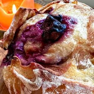 May include: Close-up of a loaf of bread with a purple filling and a dark crust. The bread has a light brown exterior with a dusting of white powder. A lit orange candle is in the background, adding warmth to the scene.