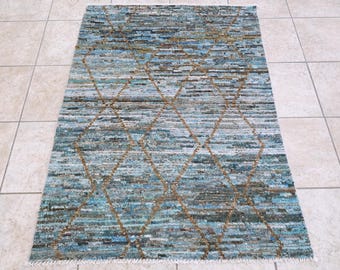 Puede incluir: Una alfombra rectangular que presenta un tejido horizontal de múltiples tonos de azul, verde azulado y marrón. La alfombra está superpuesta con un patrón de diamantes prominente y repetitivo en color beige, con flecos blancos en ambos extremos.