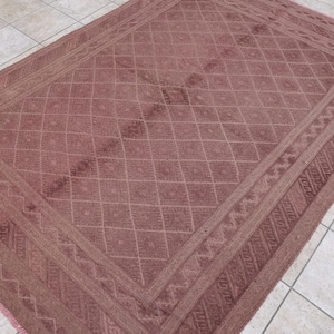 May include: A rectangular, dusty rose-colored rug with a geometric diamond pattern across the center. The rug has a detailed border with a repeating zigzag design. The rug is placed on a tiled floor.