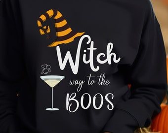 Op de afbeelding: Een zwart sweatshirt met een wit grafisch ontwerp van een heksenhoed, een martini-glas en de tekst "Witch way to the boos".