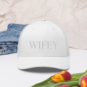 Puede incluir: Gorra de camionero blanca con la palabra "WIFEY" bordada en hilo gris claro. La gorra tiene una parte trasera de malla y una visera curva. La gorra está sobre una superficie blanca con unos vaqueros azules y flores en el fondo.