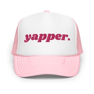May include: A pink and white trucker hat with the word "yapper." embroidered in pink on the front.