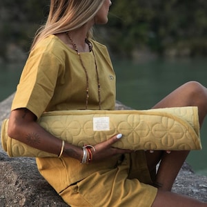 Puede incluir: Una esterilla de yoga de color amarillo mostaza con un diseño floral enrollada, sostenida por una persona con una camisa y pantalones cortos amarillos mostaza a juego. La esterilla tiene una etiqueta blanca. La persona está sentada en una roca.