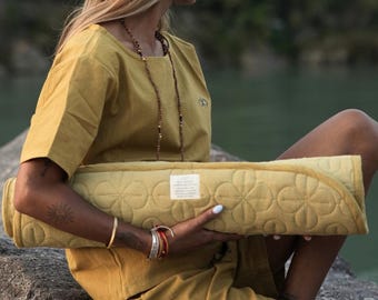May include: A mustard-yellow yoga mat with a floral pattern is rolled up and held by a person wearing a matching mustard-yellow shirt and shorts. The mat has a white label. The person is sitting on a rock.