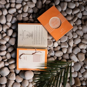 May include: An orange rectangular box with the text HEART IT OUT and KOSMOH logo, alongside an open booklet with a cream-colored page featuring text and a yoga illustration, and a separate cream-colored card with a brown string and red pattern.