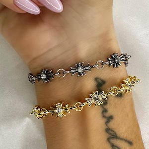 May include: Two bracelets with a floral design. One is gold-coloured, the other silver-coloured. Both are linked with small circular links. The bracelets are worn on a wrist with a tattoo.