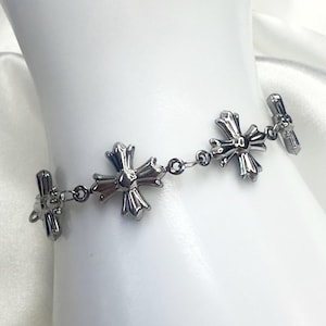 May include: A silver bracelet with three decorative cross pendants linked by small chains. The bracelet has a toggle clasp. The jewellery is shown against a white backdrop.