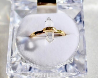 Könnte beinhalten: Ein goldener Ring mit einem klaren Quarzkristall in der Mitte. Der Ring wird in einer durchsichtigen Plastikringbox präsentiert.