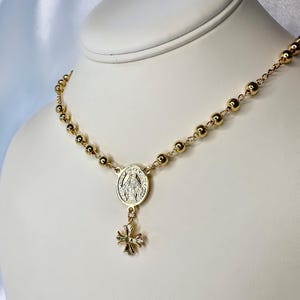 May include: Gold-toned rosary necklace with round beads and a pendant featuring a religious emblem and a small cross. The necklace is displayed on a white mannequin, showcasing its design and details.