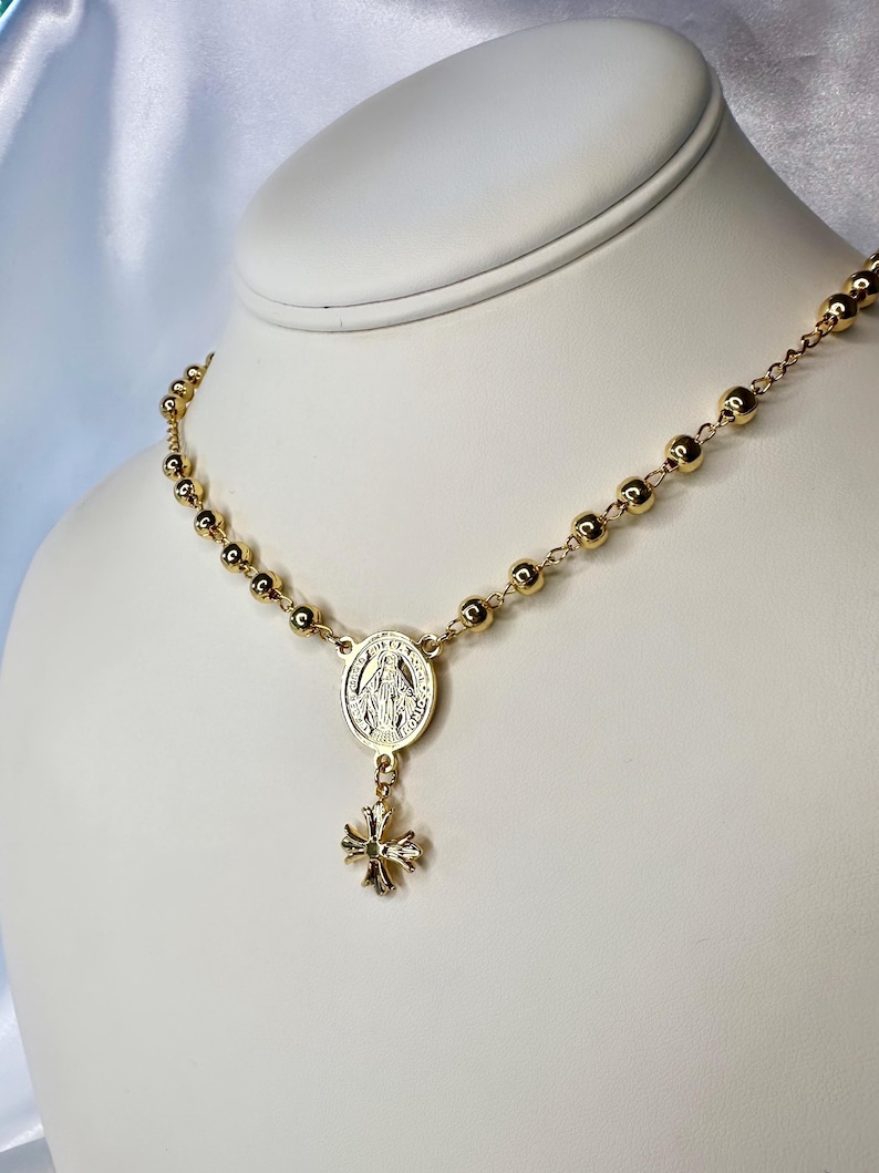 May include: Gold-toned rosary necklace with round beads and a pendant featuring a religious emblem and a small cross. The necklace is displayed on a white mannequin, showcasing its design and details.