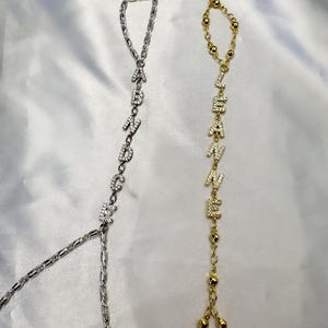 以下が含まれることがあります： 2つのハンドチェーン。1つはシルバー、もう1つはゴールドで、それぞれ異なる名前がキラキラ光る文字で綴られています。シルバーのチェーンは「ABANDONED」と綴られ、ゴールドのチェーンは「LEANNE」と綴られています。どちらのチェーンにも指輪が付いています。