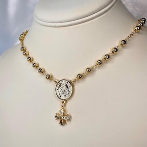 May include: Gold-toned rosary necklace featuring a central medallion with a religious figure and a decorative cross pendant. The necklace has small, round beads and is displayed on a white mannequin bust.