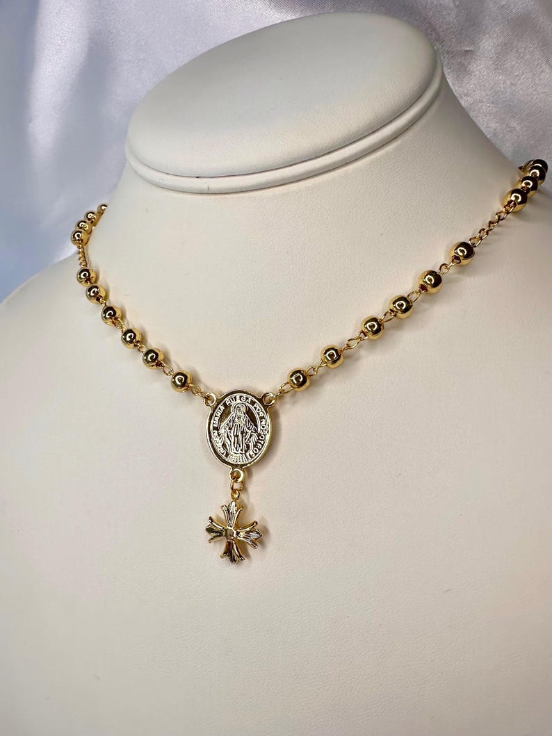May include: Gold-toned rosary necklace featuring a central medallion with a religious figure and a decorative cross pendant. The necklace has small, round beads and is displayed on a white mannequin bust.