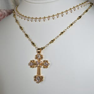 May include: A gold chain necklace with a large, ornate cross pendant featuring clear rhinestones. The necklace is layered over a delicate pearl chain with dangling pearls.