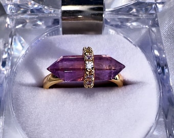 Puede incluir: Un anillo de oro con una piedra preciosa de amatista púrpura en forma de un largo cristal puntiagudo. La amatista está engastada en una banda de oro con una fila de pequeños diamantes transparentes.