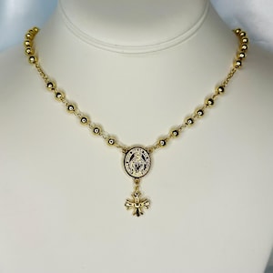 May include: Gold-toned rosary necklace featuring small, round beads and a central medallion with a religious figure. A decorative cross pendant hangs below the medallion. The necklace is displayed against a white background.