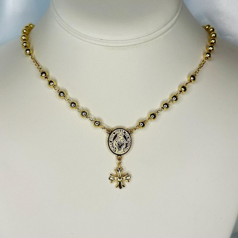 May include: Gold-toned rosary necklace featuring small, round beads and a central medallion with a religious figure. A decorative cross pendant hangs below the medallion. The necklace is displayed against a white background.