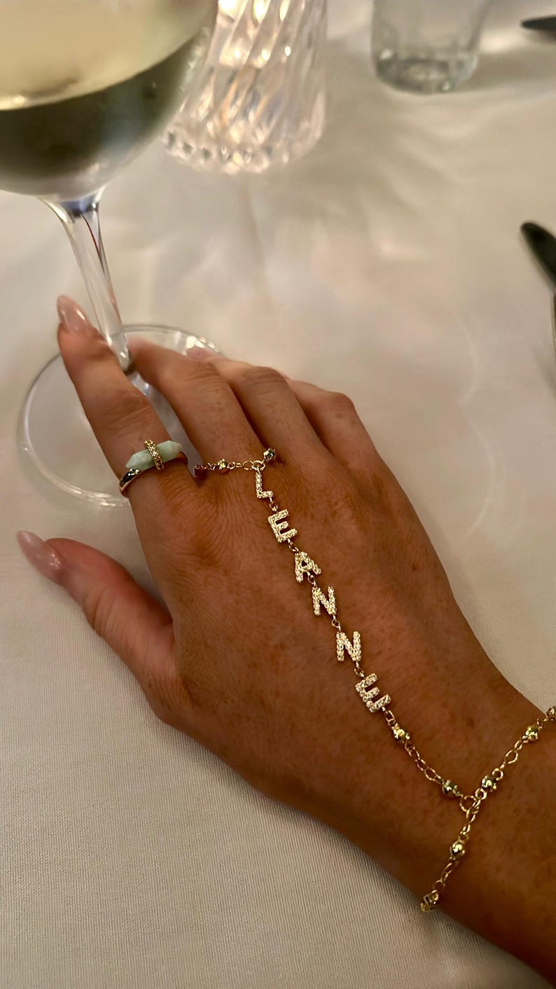 以下が含まれることがあります： 薄い緑色の石が付いたゴールドの指輪と「LEANNE」と綴られたゴールドのハンドチェーンをつけた手。ジュエリーは白いテーブルクロスの上に置かれ、背景にはワイングラスがあります。