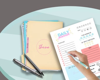 May include: A white daily planner with pink and blue accents. The planner has a to-do list, a section for tracking hydration and sunscreen application, and a notes section. The planner is on a table with a folder and pens.