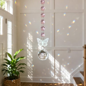 Op de afbeelding: Een hangend zonnevanger-ornament met een verticale ketting van zes gefacetteerde lichtroze kristallen kralen, een witte metalen vlinderuitsnede en een grote heldere gefacetteerde kristallen bol aan de onderkant, opgehangen aan een zilveren spiraalhaak.