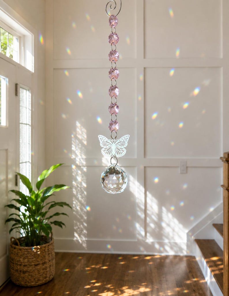 Op de afbeelding: Een hangend zonnevanger-ornament met een verticale ketting van zes gefacetteerde lichtroze kristallen kralen, een witte metalen vlinderuitsnede en een grote heldere gefacetteerde kristallen bol aan de onderkant, opgehangen aan een zilveren spiraalhaak.