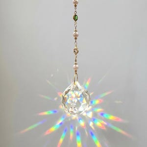 May include: A hanging crystal suncatcher featuring a faceted clear glass teardrop prism that refracts light into a vibrant rainbow starburst pattern. The prism is suspended by a gold-toned metal chain accented with small white pearls and green beads.