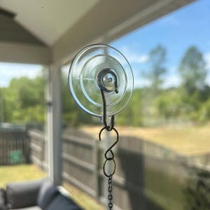 Könnte beinhalten: Ein transparenter Saugnapf mit Metallhaken und Kette, zum Aufhängen von Gegenständen an einem Fenster oder einer glatten Oberfläche. Der Saugnapf ist rund und transparent, mit einer Metallschlaufe zur Befestigung des Hakens. Die Kette besteht aus kleinen, verbundenen Metallstücken.