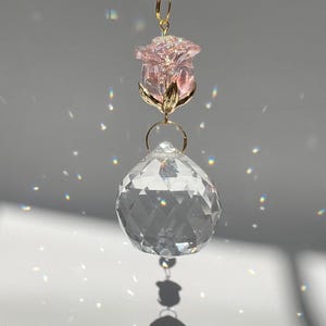 May include: A decorative crystal suncatcher with a faceted clear crystal ball, a pink rose, and a smaller clear crystal at the top. The rose has gold-colored accents. The suncatcher hangs from a gold-colored chain, creating a play of light and shadows.