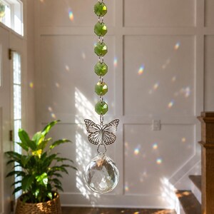 Op de afbeelding: Een hangende zonnevanger met een verticale streng van gefacetteerde limoengroene kralen verbonden door zilverkleurige metalen ringen. Een zilverkleurige vlinderbedel hangt onder de kralen, gevolgd door een grote, heldere, gefacetteerde kristallen bol.