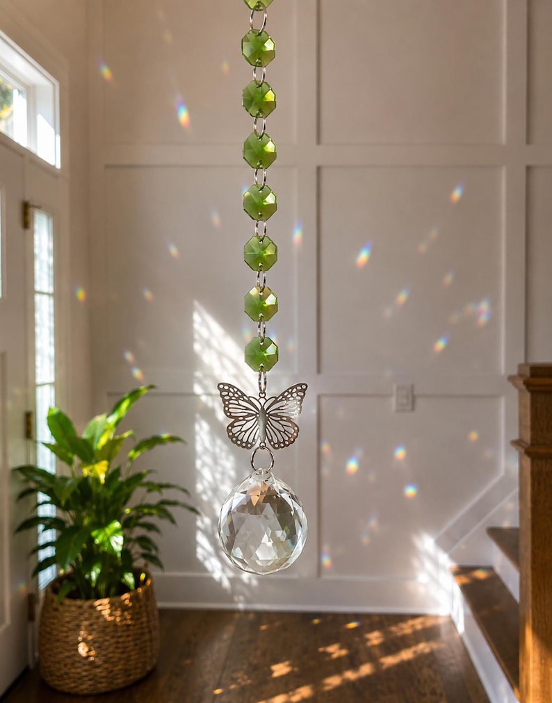 Op de afbeelding: Een hangende zonnevanger met een verticale streng van gefacetteerde limoengroene kralen verbonden door zilverkleurige metalen ringen. Een zilverkleurige vlinderbedel hangt onder de kralen, gevolgd door een grote, heldere, gefacetteerde kristallen bol.