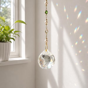 May include: A hanging crystal suncatcher featuring a faceted clear glass sphere suspended from a gold-toned chain. The chain is embellished with alternating white pearls, small clear crystals, and two colored beads in teal and green hues.