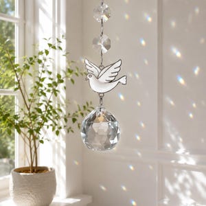 May include: A hanging suncatcher ornament featuring three clear faceted crystal beads at the top, a white enameled dove charm in the center, and a large clear faceted crystal sphere at the bottom, all connected by silver-toned metal links.
