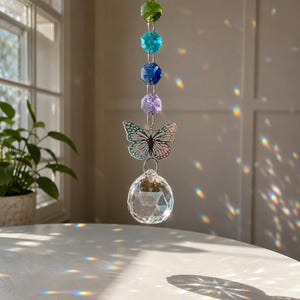 Op de afbeelding: Een hangende zonnevanger met een verticale ketting van vier gefacetteerde kralen in groen, turquoise, donkerblauw en lichtpaars. Onder de kralen hangt een zilverkleurige metalen vlinderbedel aan een helder, bolvormig gefacetteerd kristallen prisma.